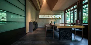 A modern dining room with a long table, wooden floor, and large glass windows showcasing a green exterior.
