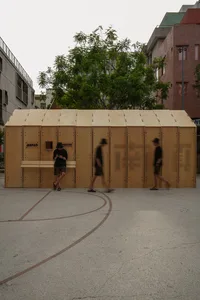 A minimalist wooden shelter in an urban setting, with people walking past.