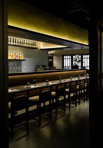 A modern restaurant bar with wooden flooring, tables, and chairs, illuminated by soft lighting.
