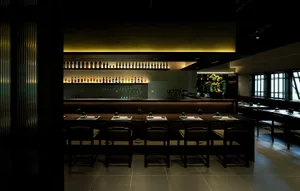 A modern restaurant interior with sleek wooden tables, black chairs, and warm yellow lighting.
