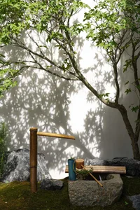 A serene Japanese-style garden water feature with a wooden spout and bamboo pipe, surrounded by rocks and greenery.