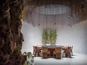 A minimalist dining room with a round wooden table and chairs, set against a textured white wall with small green plants and 