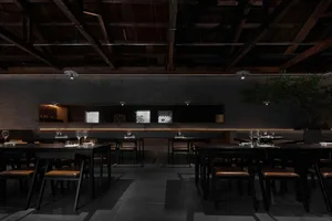 A minimalist restaurant interior featuring concrete walls and dark tables and chairs.