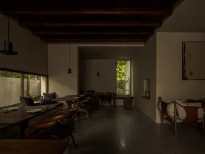 Indoors, a minimalist restaurant with wooden tables and concrete floors, featuring tall ceilings with wooden beams, large win