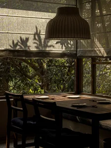 A modern dining room featuring a wooden table with plates and chopsticks, surrounded by fabric seating and glass windows with