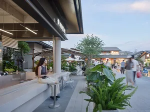 A person sitting on a modern, concrete outdoor bench in front of a restaurant with green foliage and pedestrians in the backg
