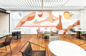 An indoor café with wooden flooring, featuring abstract art on the wall, modern seating, and multiple white tables.