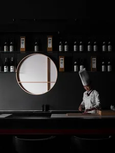 A minimalist restaurant interior with dark walls and a white table, featuring a chef preparing food.