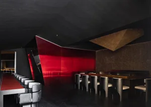A sleek modern restaurant bar with red leather stools, black concrete walls, and a geometric light fixture.