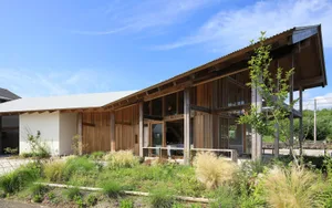 A modern cabin with a wooden exterior, green shrubs in the forground, and a clear blue sky above.