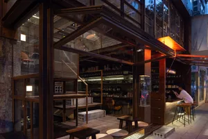 An industrial-style restaurant interior featuring wooden tables, glass shelves, and concrete surfaces.