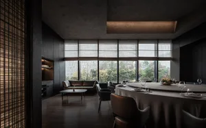 Modern restaurant dining room with round table, chairs, and large windows overlooking greenery.