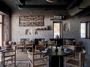 A cozy restaurant interior made of clay, wood, and concrete materials with woven chairs and round tables.