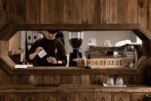 A barista working behind the counter of a coffee shop with wooden materials and a clock showing dates.