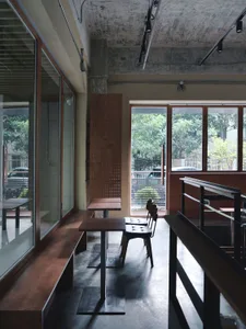 A modern cafe interior with wooden tables and chairs, concrete flooring, and large windows showing greenery outside.