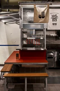 A minimalist restaurant interior with a metal food counter, wooden benches, and Korean signage.