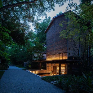 A modern house with brick and concrete exterior surrounded by lush greenery and illuminated by warm lights.