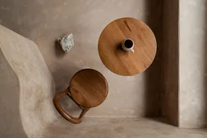 A modern wooden table with a small ceramic mug and a wooden stool, alongside a rock on a textured beige wall.