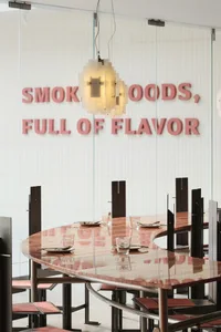 Modern restaurant dining room with wooden tables, concrete walls, and the words 'Smoked Goods, Full of Flavor' on the wall.