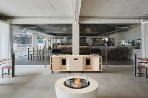 A modern restaurant bar with a concrete floor, industrial-style furniture, and a central fire pit.