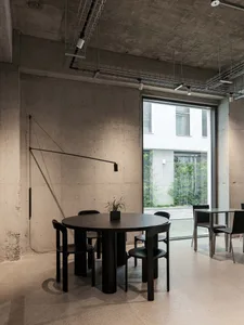 Industrial-style cafe interior with concrete walls and ceiling, featuring round tables and black chairs.