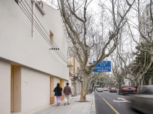 A minimalist street scene featuring concrete buildings and bare trees on a cloudy day, with people walking on the sidewalk.