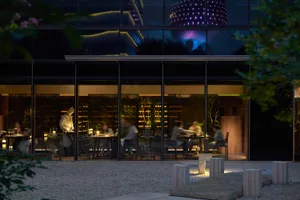 A modern restaurant exterior at night with a glass facade reflecting city lights and a chef working inside.