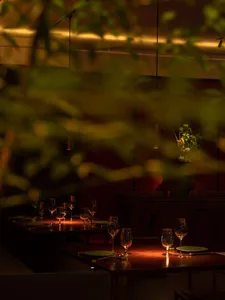 Modern restaurant interior with dark lighting and wooden tables and chairs, glassware, and green plates.