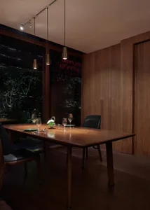 Modern dining room with wooden table, chairs, and pendant lights, featuring a view of greenery outside through large windows.