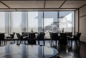 A modern restaurant interior with black tables, chairs, and floor, large windows with sheer curtains, and a view of a tree an