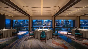 A modern restaurant with blue upholstered chairs, glass windows overlooking a cityscape, and a carpeted floor.