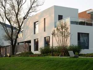 Modern exterior of a residential home featuring white concrete walls, glass window panels, and surrounding foliage.