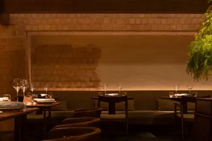 A modern restaurant interior featuring wooden tables and chairs, with a brick wall and green foliage.