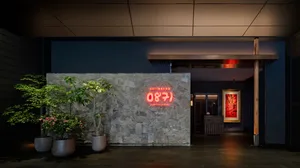 A modern restaurant interior with concrete walls, plants, and a neon sign.