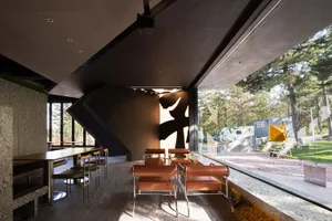 Modern coffee shop interior with black concrete ground and glass walls showing outdoor seating area with trees.