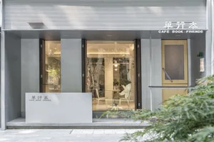 Modern cafe with glass frontage, seating area inside, and greenery outside.