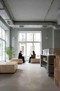 Two people sitting on wooden benches in a modern cafe interior with concrete walls and floor, and floor-to-ceiling windows ov
