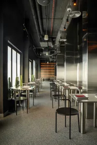 An empty, industrial-style restaurant with long tables, black chairs, and glass partitions.
