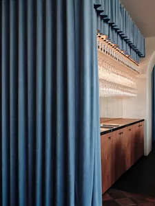 A modern cafe bar featuring a blue velvet curtain, a wooden bar counter, and a metal circular ceiling light fixture.