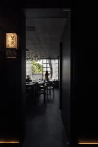 A minimalist dining room with concrete walls and floor, featuring a round table and chairs, and a person adjusting curtains.