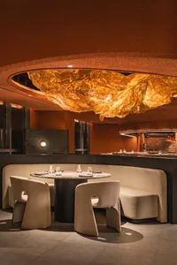 A minimalist restaurant interior with circular leather seating, a round table, and a large, illuminated chandelier hanging fr