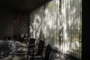 A modern restaurant dining area with sheer curtains, glass walls, and minimalist black chairs.