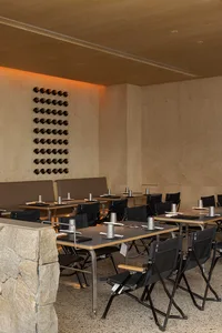 A modern restaurant interior with wooden tables and chairs, featuring a stone pillar and a wine rack on the wall.