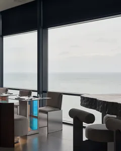 Modern restaurant interior with metallic tables, stone benches, and upholstered seating overlooking the ocean.