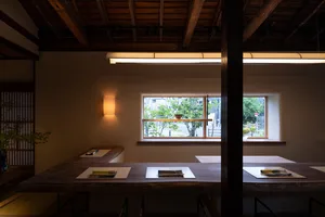 A minimalist restaurant interior with wooden elements, featuring a counter and windows with outdoor views.