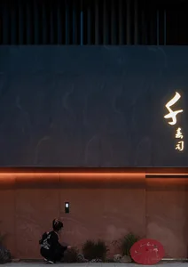 Person wearing a black kimono and a red umbrella standing in front of a modern building with brick facade and neon lights.