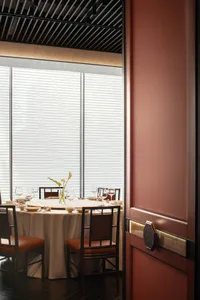 A minimalist restaurant interior featuring white blinds, wooden chairs, fabric tablecloth, and glass drinkware.