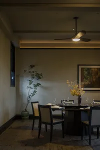 A minimalist dining room featuring natural wood elements, under warm lighting with a ceiling fan and a framed artwork.