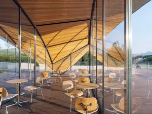 A modern cafe interior with wooden tables and chairs, large windows, and a wooden roof with geometric shapes.