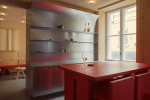 A minimalist dining room with a red wooden table and chairs, featuring a metallic shelving unit and a window with a view of a